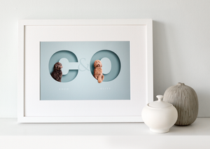 white wood framed light blue picture of an apricot coloured cockapoo and a chocolate brown cockapoo. each dog is sitting in the initial letter of their name that has a cut out of paper design effect and their full name is in an elegant serif typeface below